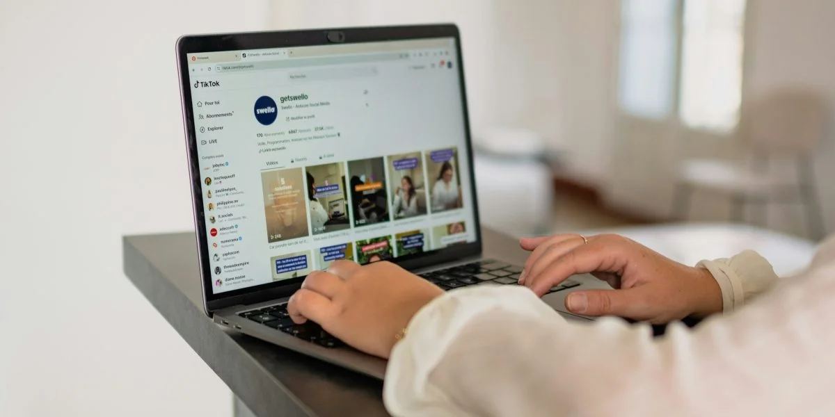 A person typing on a laptop, viewing a social media platform with posts and a profile page open on the screen